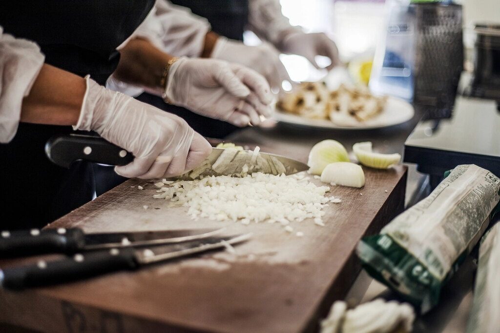 Des mains gantées taillent des oignons sur une planche, en cuisine professionnelle.
